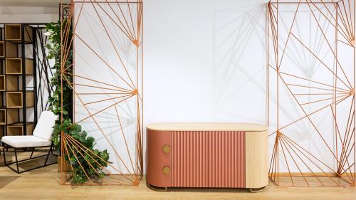 T-DESIGN Sideboard in Copper Tones – A Harmonious Interior with REZES BANDA Room Dividers The image showcases a compact T-DESIGN sideboard featuring a distinctive copper-blush front that brings warmth and elegance to the space. The finely milled fluted surface is complemented by natural oak accents and brass details, creating a timeless yet contemporary piece of furniture. The sideboard is framed by decorative REZES BANDA room dividers, which serve not only as functional space separators but also as striking design elements that define the interior.